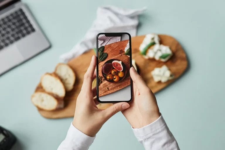 Overhead shot of a person taking a picture of a cheese, fruit, and bread board with their smartphone.
