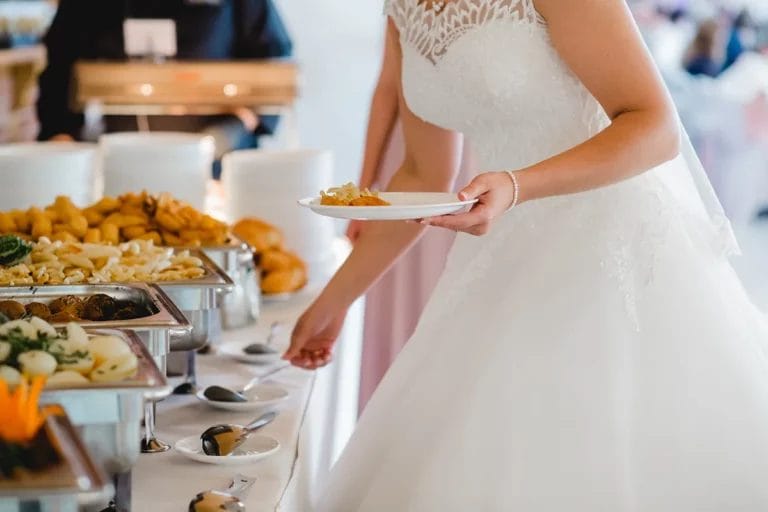 A bride fills her plate with food from a catering buffet table at a wedding.