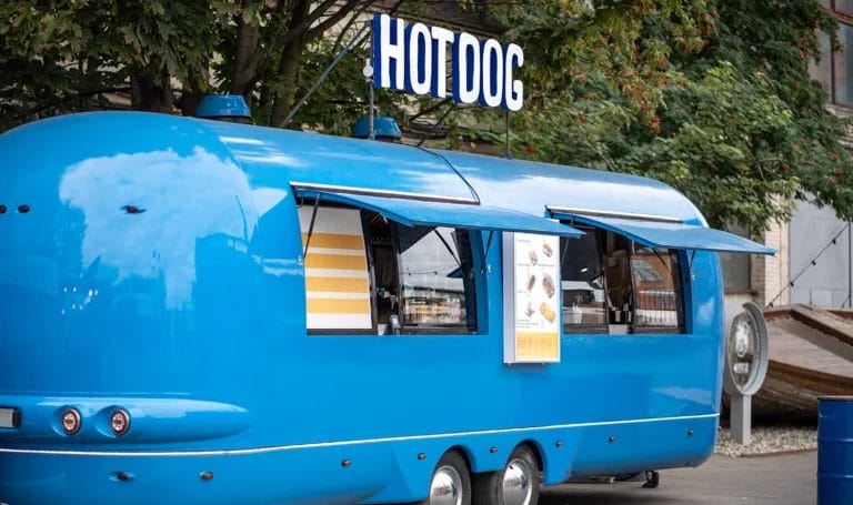 A blue food truck with the words "Hot Dog" on top sits on an empty street waiting for customers.