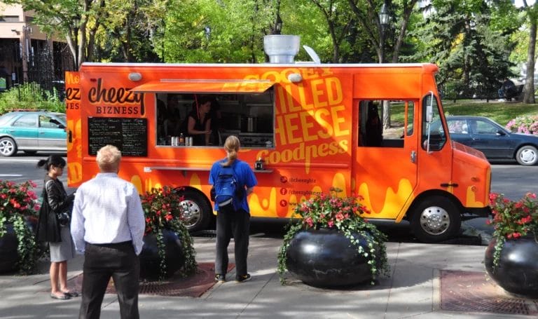 The Cheezy Bizness food truck is parked outside in a shady business park, serving customers grilled cheese sandwiches.