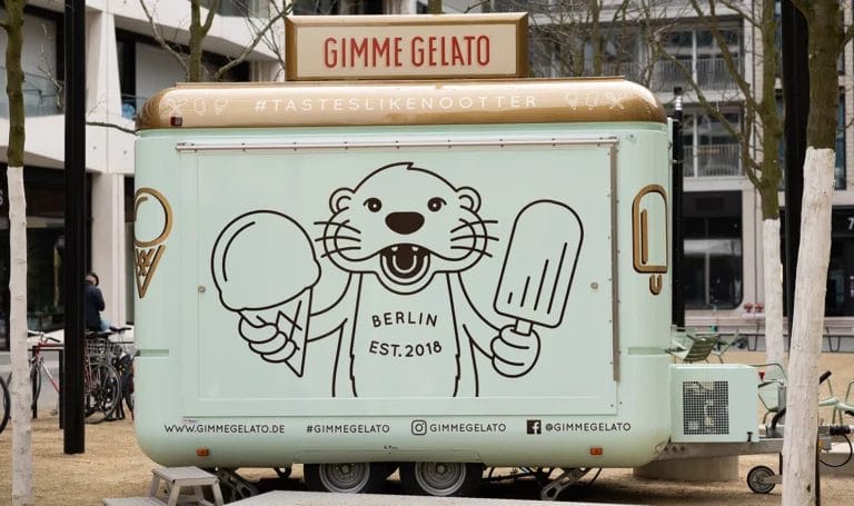 A mint green colored food trailer is parked and closed for the day in an empty public space. It's branding reads, "Gimme Gelato," with the hashtag taste like no other and a large logo of an otter eating ice cream adorning the front of the trailer.