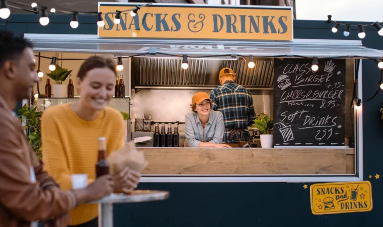 A food truck owner leans on the edge of her counter and smiles out at her happy customers enjoying their food from her truck.