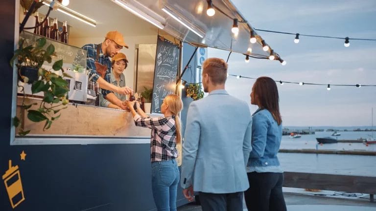 Food truck workers hand out orders from the window at dusk, with hanging string lights above and a harbor in the background.