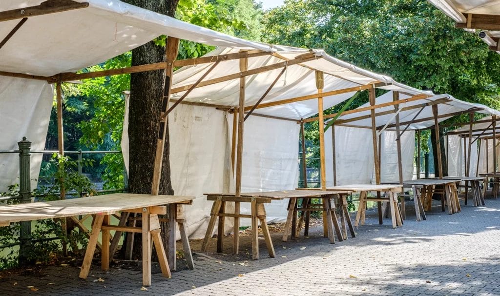 Empty wooden vendor tables with fabric canopies line an open-air marketplace before a festival.