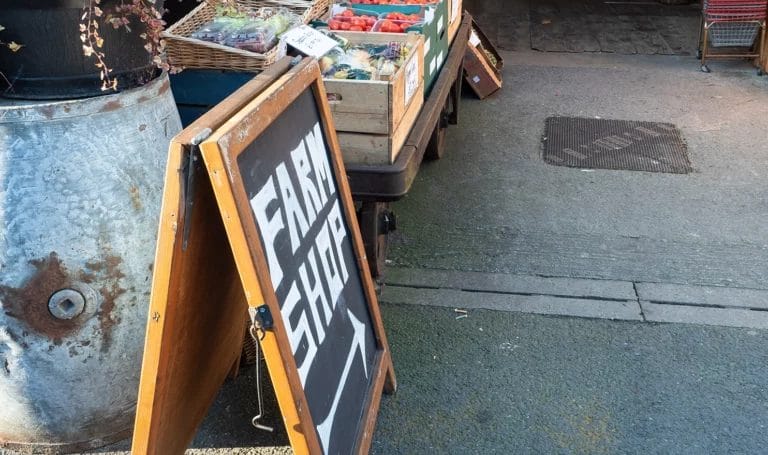 An a-framesign that reads "Farm Shop" is propped up next to a booth with fresh product at an outdoor market.