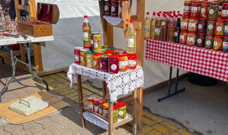 Colorful jars of handmade jams and sauces on display in a vendor booth at an outdoor farmers market. The jars are on tables with lace and red gingham tablecloths.