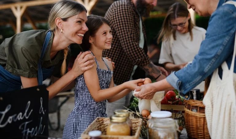 Happy woman buying organic eggs from a mother and daughter duo at an outdoors local farmers market.