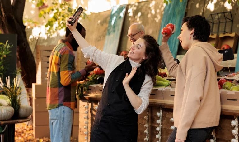 Two shoppers smile and pose with their smart phone as they take a selfie in front of a farmers market booth.
