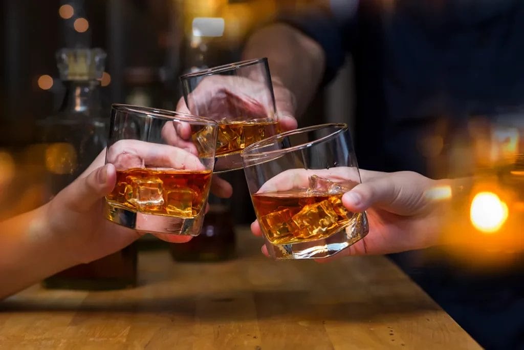 Three people clink glasses of whiskey together over a wooden table.