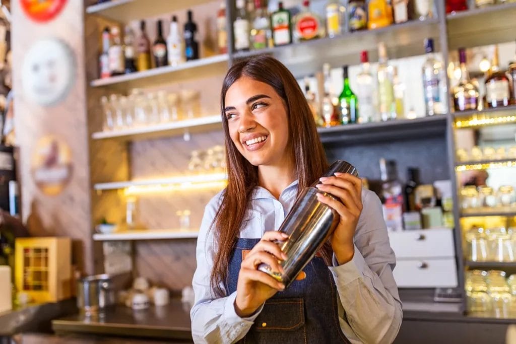 A smiling female bartender with long brown hair holds a cocktail shaker behind a bar.