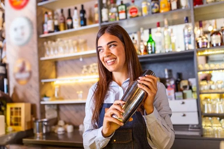 A smiling female bartender with long brown hair holds a cocktail shaker behind a bar.