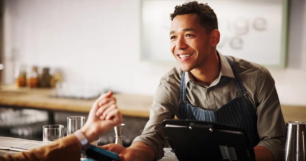 A bartender wearing a gray button-up shirt and denim apron taps a customer's smartwatch for payment while behind the bar terminal.