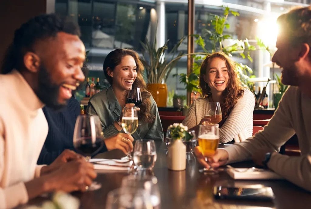 A group of people laughing while drinking glasses of wine at a table with a glass window and plants in the background.