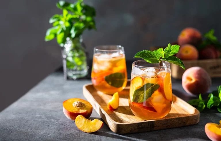 A pair of peach tea cocktails sits on a tray with cut and whole peaches and mint sprigs on a gray table.