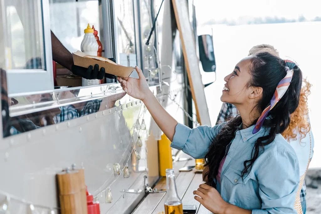 A customer smiles as an employee inside a food truck hands her a cardboard container with food.