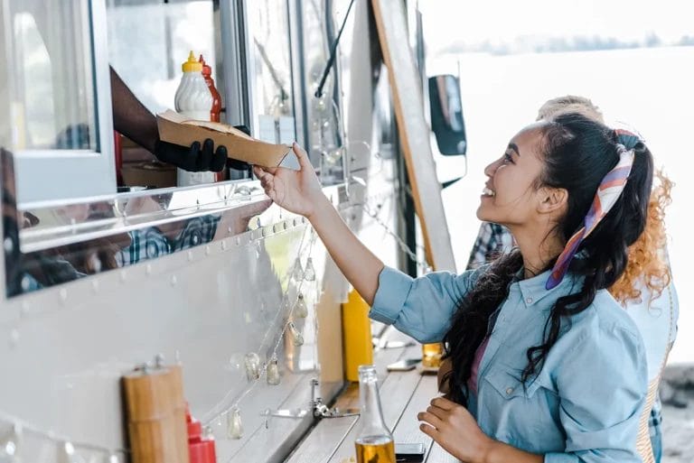 A customer smiles as an employee inside a food truck hands her a cardboard container with food.