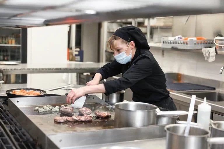 A chef wearing a protective face mask cooks meat in a commercial kitchen.