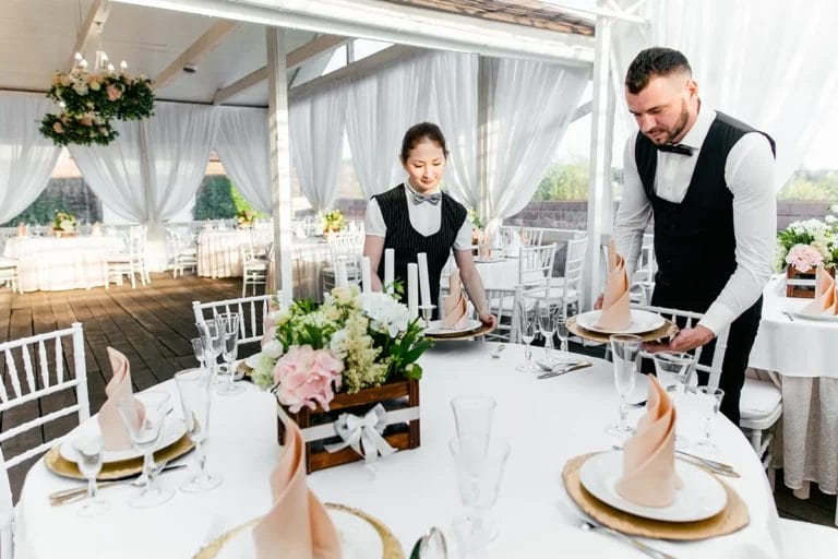 Two catering servers place plates with decorative napkins on a table at a wedding reception.