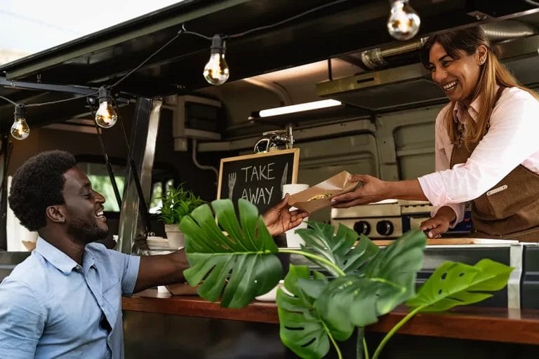 A food truck worker in a brown apron hands a takeout basket of food to a customer.
