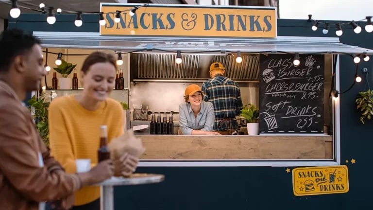 Black and yellow snacks and drinks food truck with a smiling employee and customers eating in the foreground.