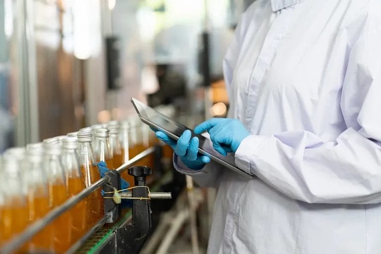 A close-up shot of an employee in a food manufacturing plant holding a tablet and inspecting a conveyor belt with bottled beverages.