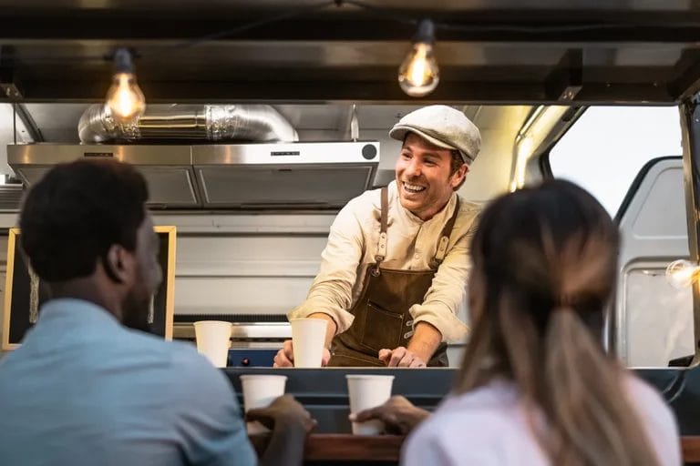 A man wearing a gray cap and brown apron smiles while handing beverages to two customers at his food truck.