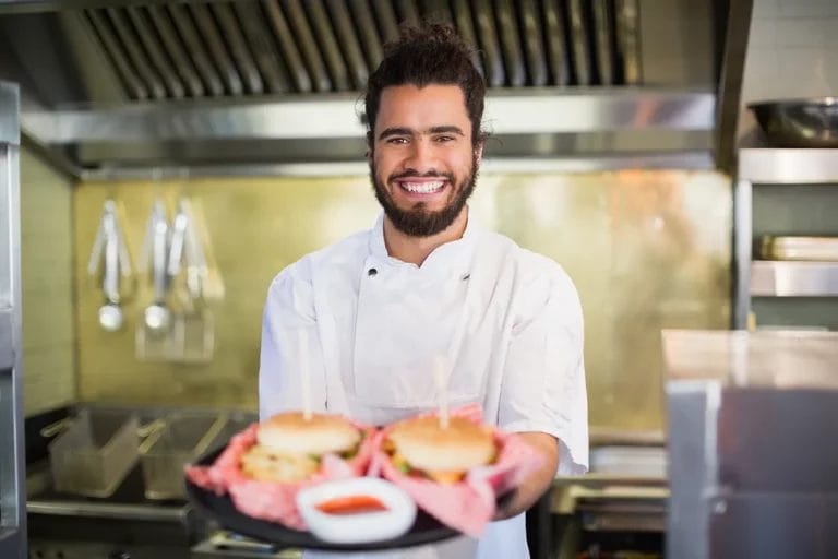 A male chef inside a stainless steel commercial kitchen holds a plate with two burgers while smiling at the camera.