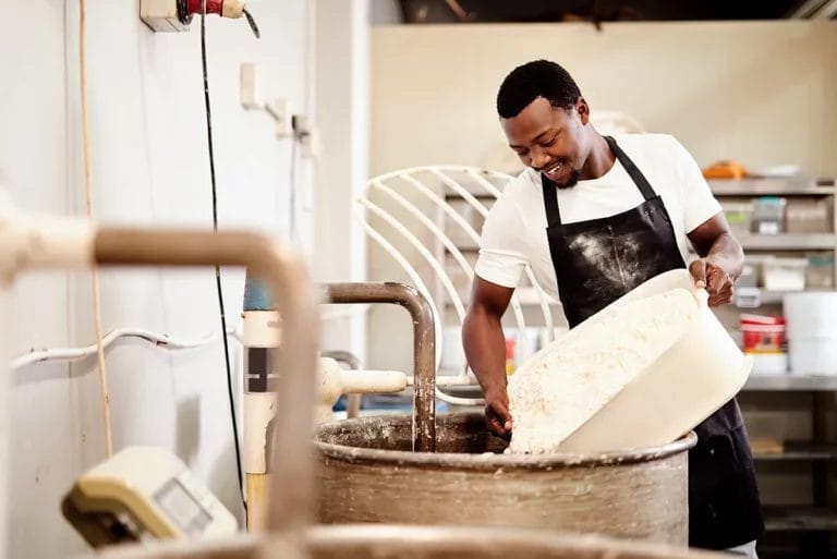 A male bakery employee wearing a white shirt and black apron pours a container of flour into an industrial stand mixer while smiling.