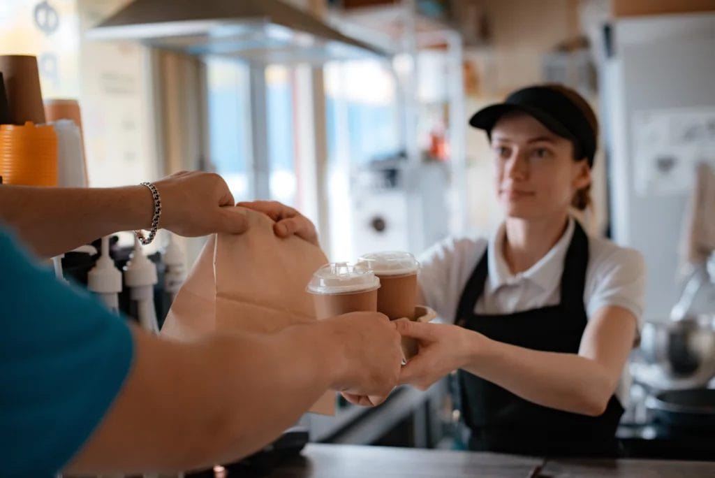 An employee in a black hat and apron hands two drinks and a bag of food to a customer over a restaurant counter.