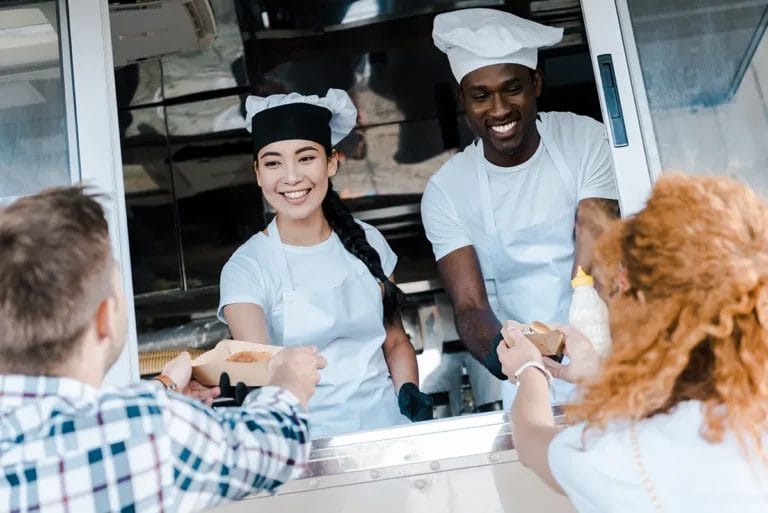 Two food truck employees in white aprons and chef hats serve food to two customers out of the service window.