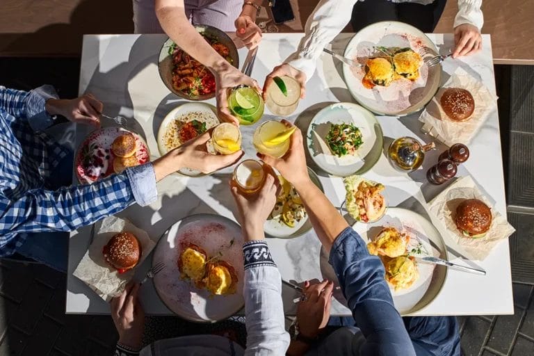 Bird's eye view of a group of friends clinking cocktail glasses over a restaurant table full of food.