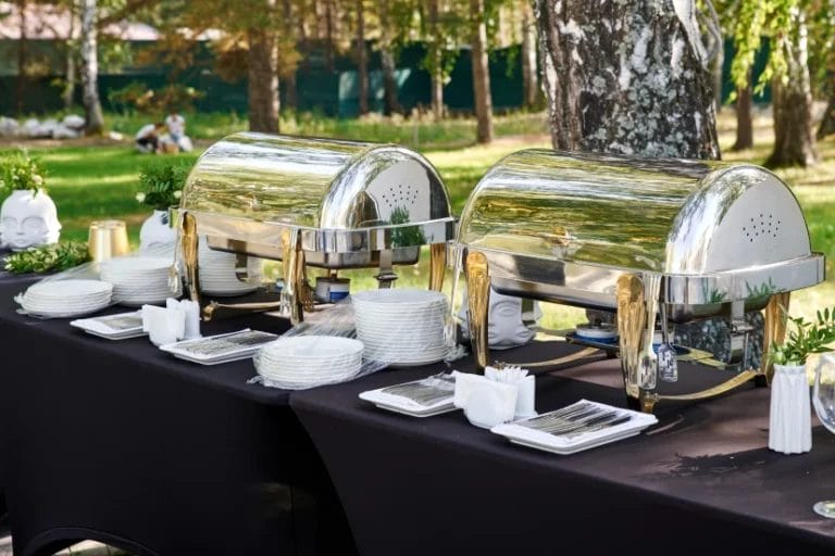 catered outdoor banquet table with chafing dishes, cutlery, and plates